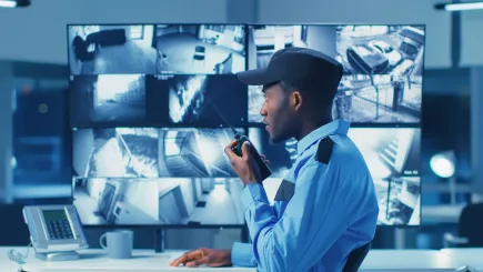 A uniformed security officer using a radio while monitoring multiple CCTV screens in a security operations centre, representing TrustForce Security's 24/7 alarm monitoring and intervention services in Qatar.