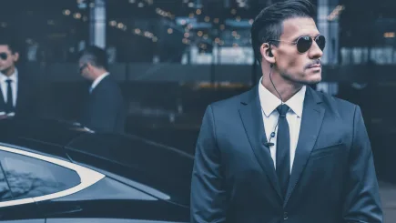 A suited close protection officer with earpiece standing beside a luxury vehicle, with a second officer visible in the background, illustrating TrustForce Security's professional executive protection services in Qatar.