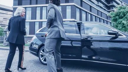 A suited protection officer opening the door of a luxury black saloon car for a female executive outside a modern commercial building, representing TrustForce Security's VIP escort and secured ground transportation services in Qatar.