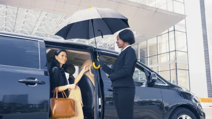 A female protection officer holding an umbrella and assisting a VIP client stepping out of a luxury black people carrier outside a modern building, representing TrustForce Security's secured ground transportation and VIP escort services in Qatar.