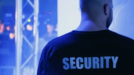 A security officer in a black uniform with "Security" printed on the back standing watch at a live event venue, representing TrustForce Security's professional event access control and crowd management services in Qatar.