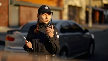 A female security officer in uniform using a radio and holding a tablet while on duty outside a commercial building, representing TrustForce Security's professional manned guarding and building security services across Qatar.