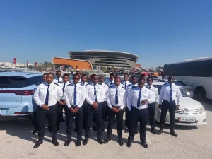 Group of uniformed security personnel posing for a photo in front of a modern stadium structure in Qatar with cars parked around them.
